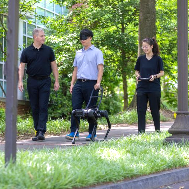 three people walkingthe man in the middle has a blindfold on and is being led by a robot dog