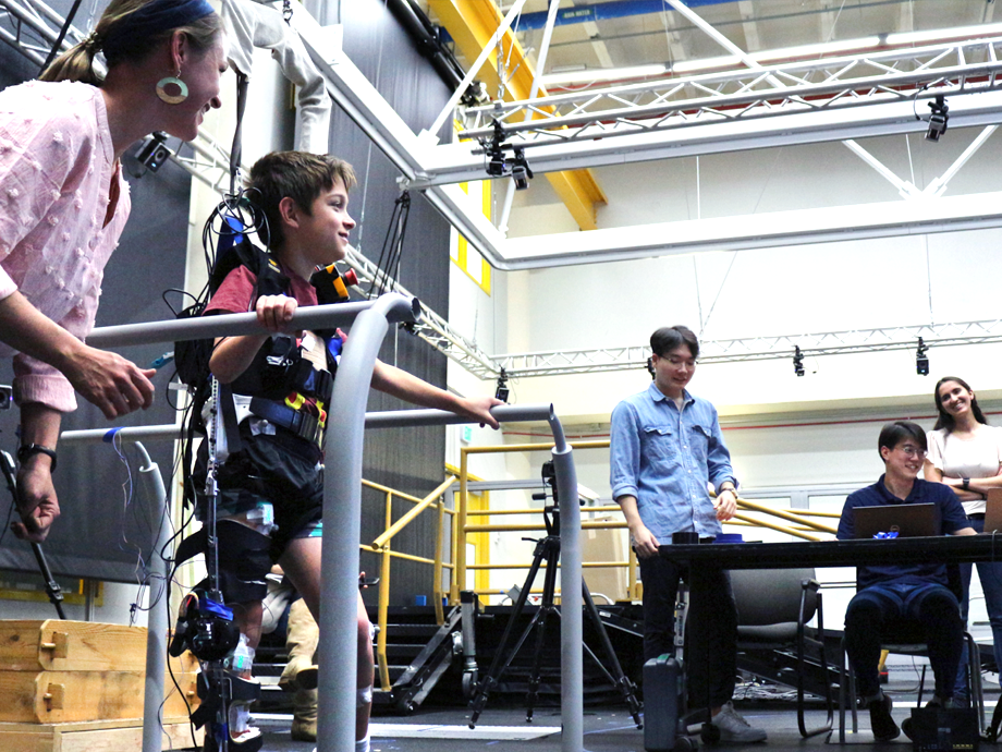 group of students and research observing a adelescent on a treadmill with a harness on connected to machines and computers
