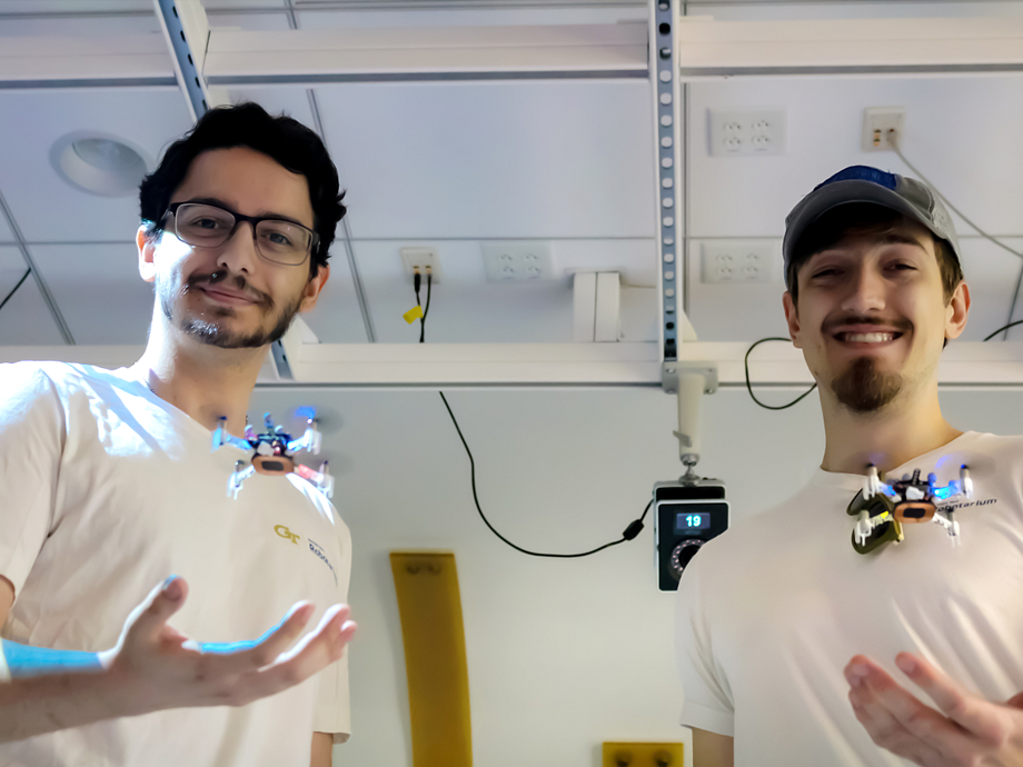 two students standing facing you, each student has a small drone flying over the hand that is extend out and palm up