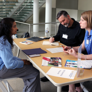 Two people at a table presenting information to a single person on the other side of the table